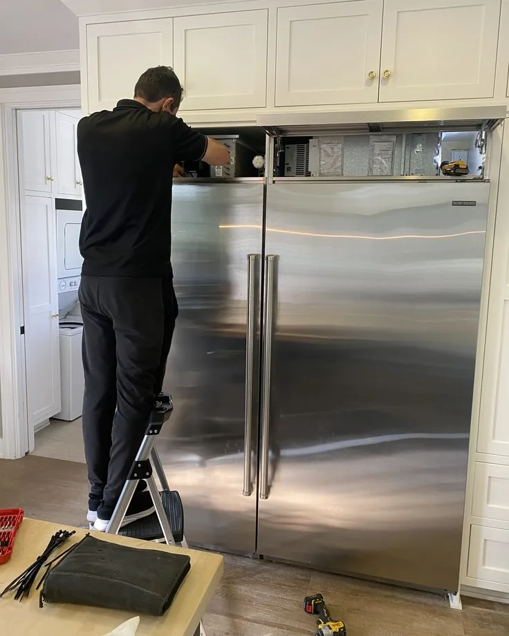 Rapid Solutions technician on a step ladder servicing the upper unit of a built-in Sub-Zero refrigerator, tools laid out nearby