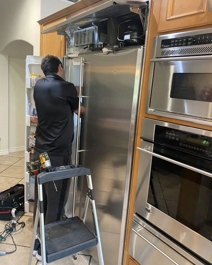 Technician servicing the condenser unit above an open built-in refrigerator, with a cordless drill and tool bag beside him